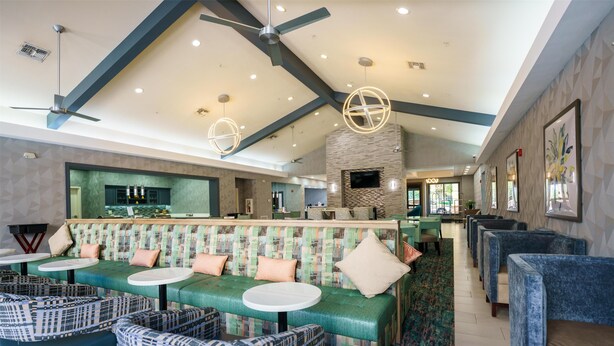A modern hotel lobby with a high ceiling, a central seating area with a green sofa and patterned chairs, a fireplace, and a TV mounted on the wall. - Homewood Suites Houston West Energy Corridor (Houston)