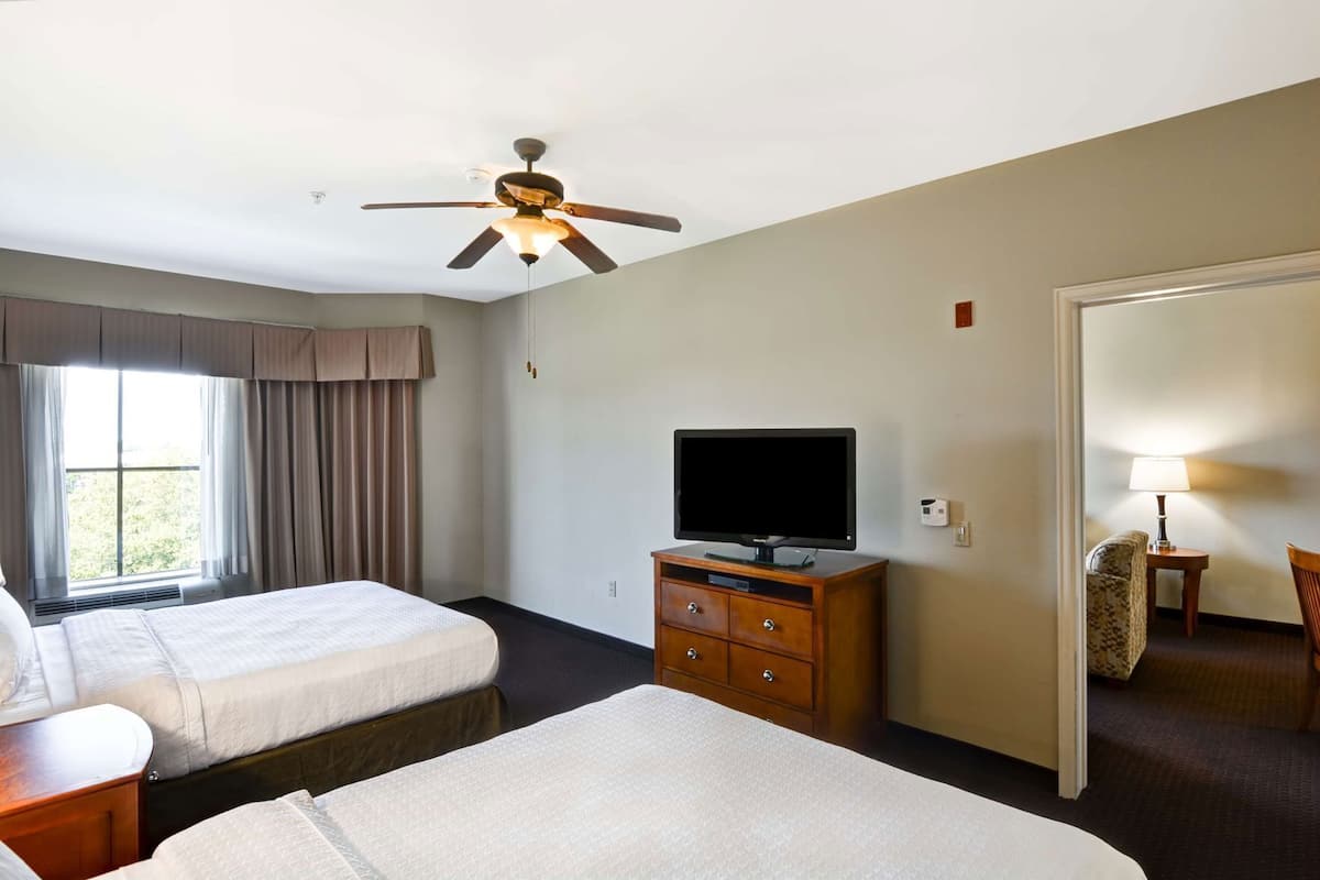 A hotel room with two beds, a television on a wooden dresser, a ceiling fan, and a window with curtains.