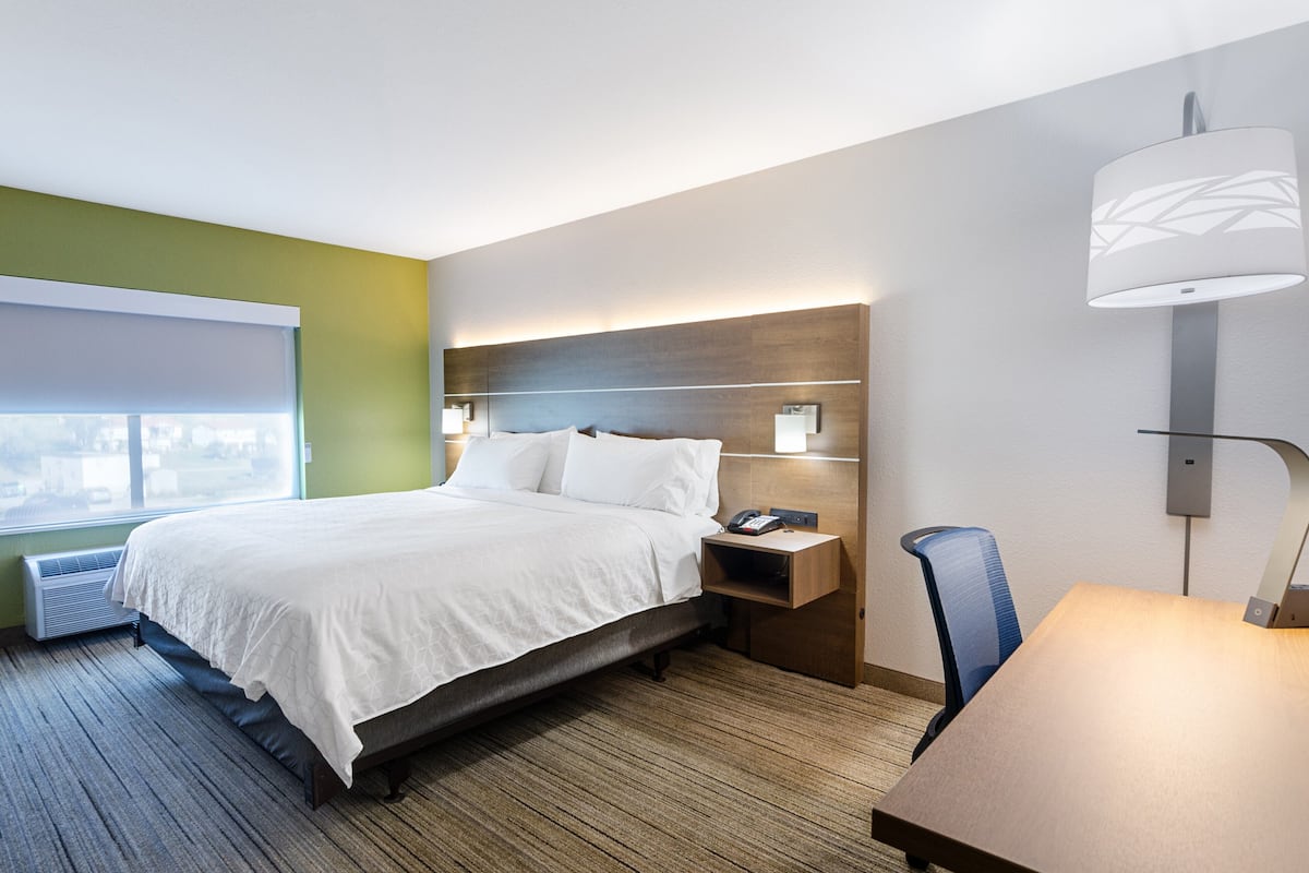 A hotel room with a bed, bedside table, lamp, and a view of the city through the window.