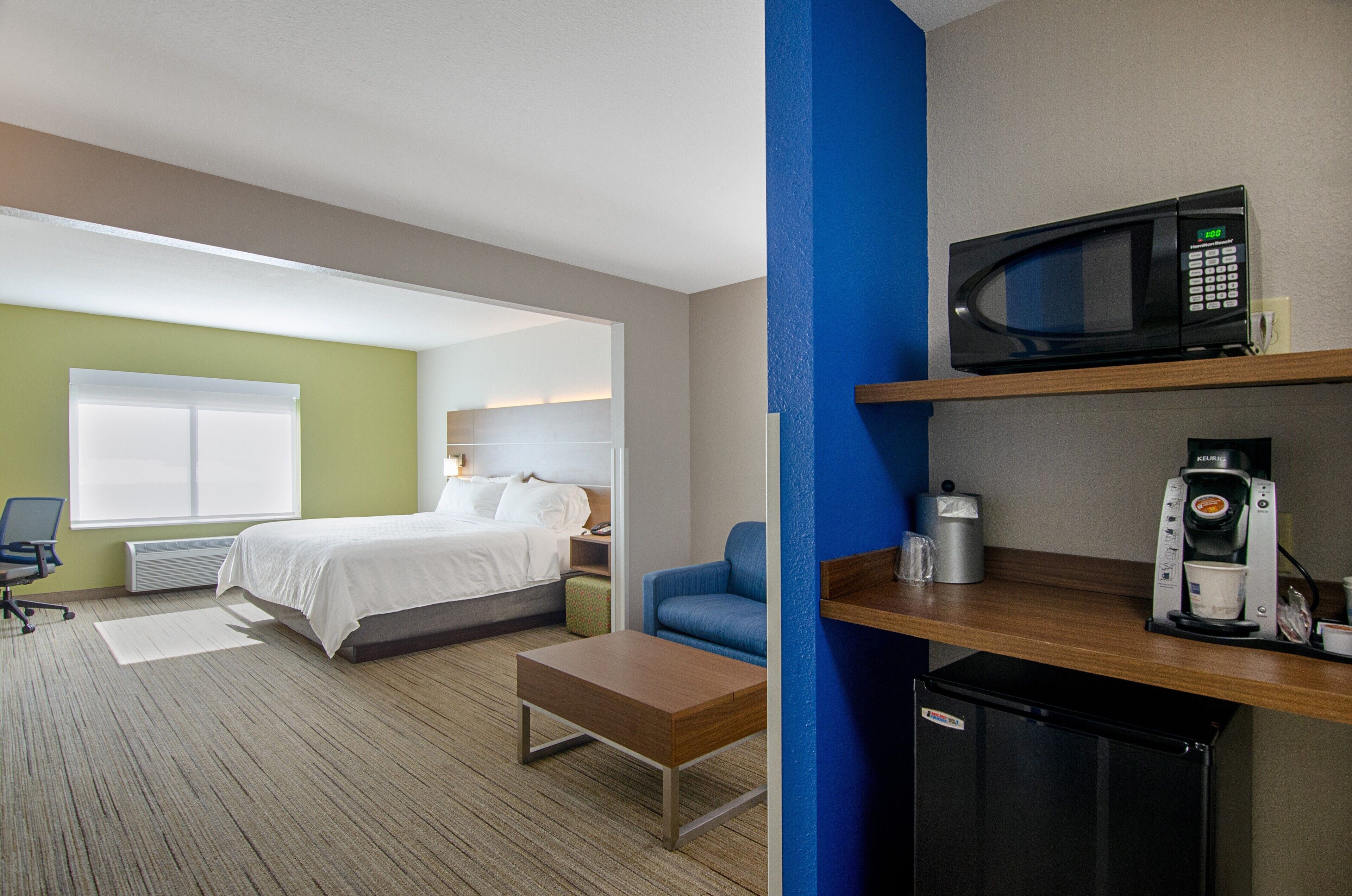 Habitación de hotel con cama, escritorio, silla, microondas, cafetera y refrigerador pequeño.
