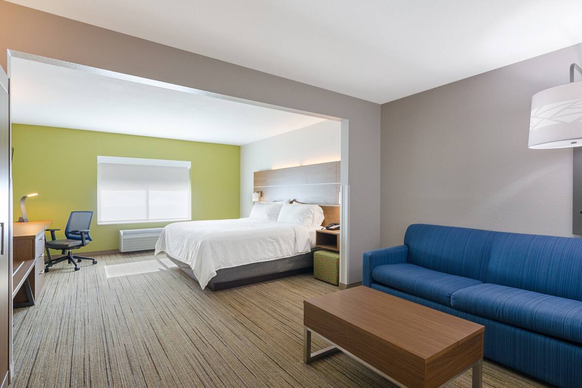 A hotel room with a bed, a blue sofa, a wooden coffee table, a desk with a chair, and a window with blinds.