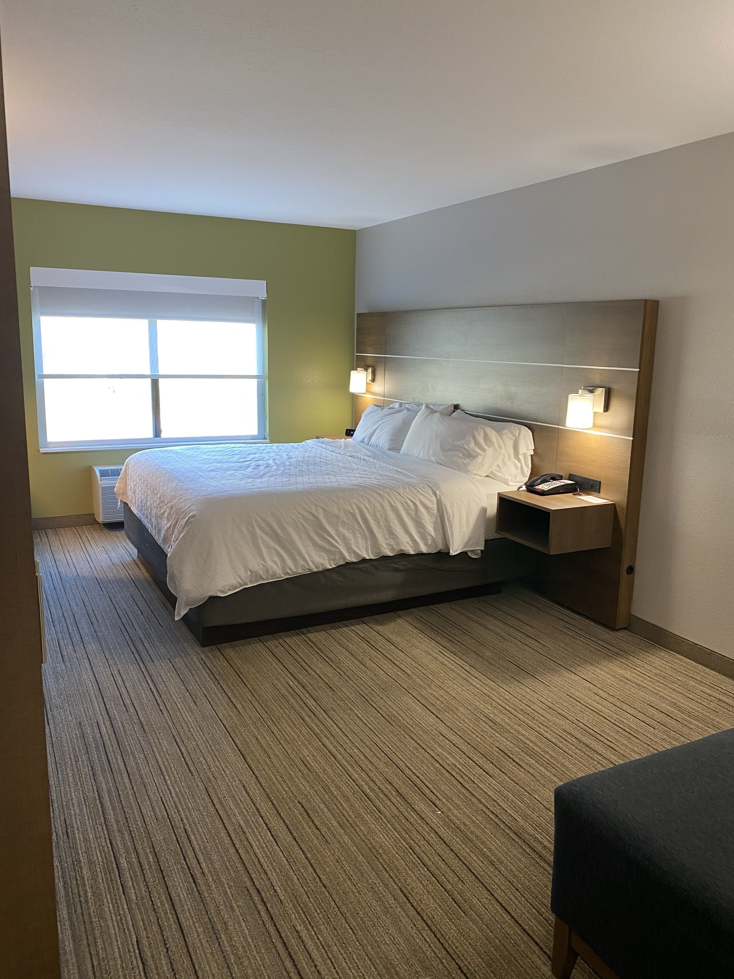 A hotel room with a large bed, a nightstand, a wall-mounted lamp, and a window with blinds.