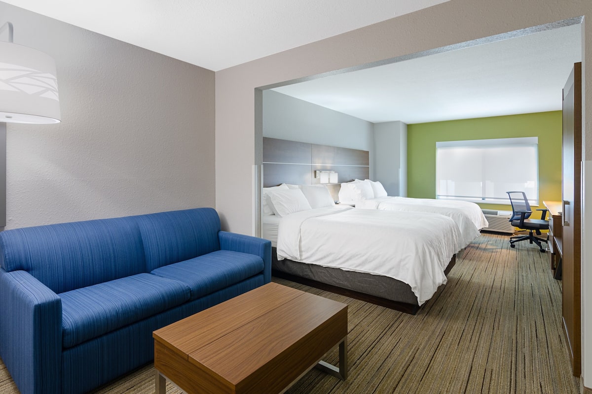 A hotel room with a blue sofa, a wooden coffee table, two beds with white bedding, a desk with a chair, and a large window with blinds.
