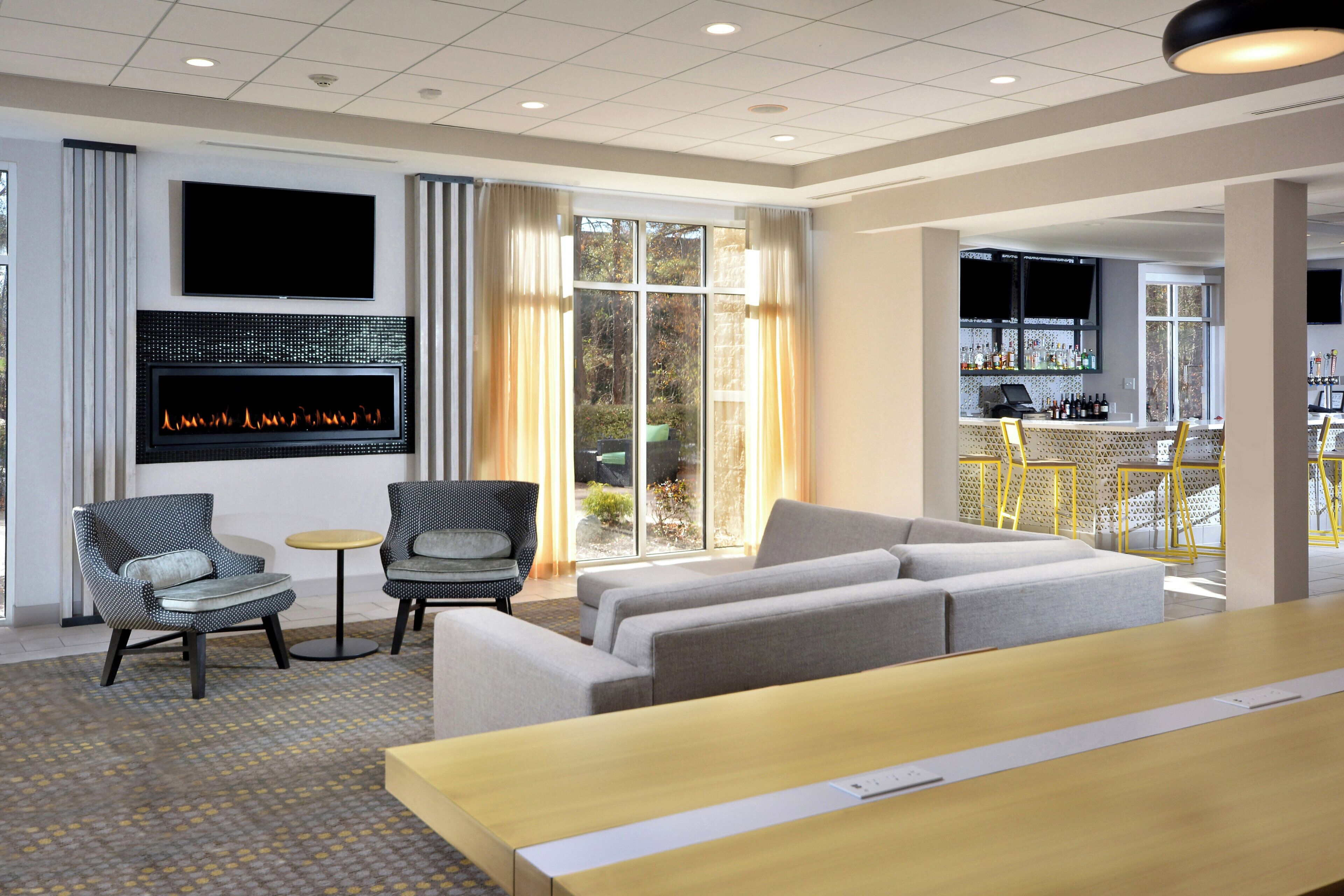 A modern hotel lobby with a seating area, a fireplace, a bar, and large windows.