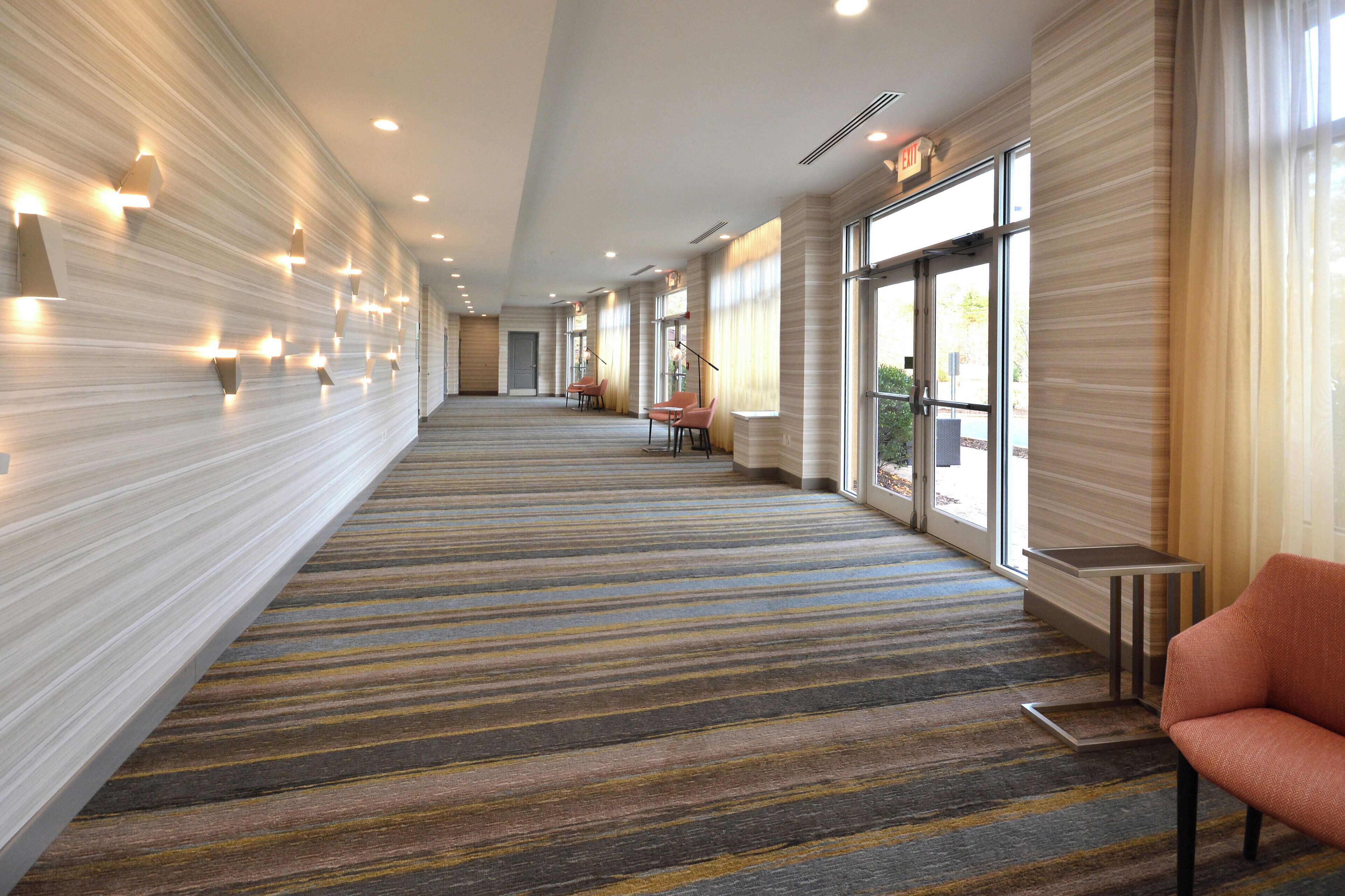 A hotel corridor with a striped carpet, wall-mounted lights, and a seating area with chairs.