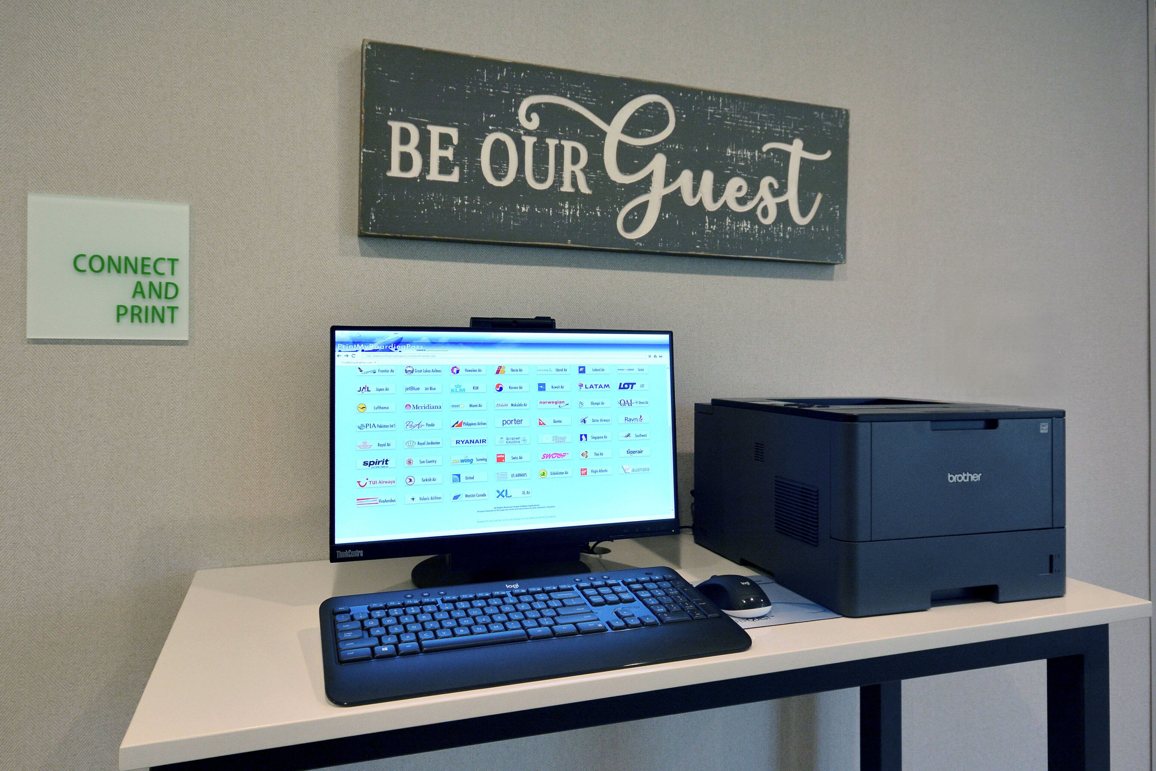 A desk with a computer, keyboard, and printer, a wall sign saying 'BE OUR GUEST,' and a sign that says 'CONNECT AND PRINT.'