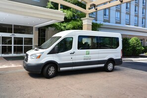 Un fourgon blanc est garé devant un immeuble orné d’une enseigne « Holiday Inn », avec de grandes fenêtres et une entrée abritée.