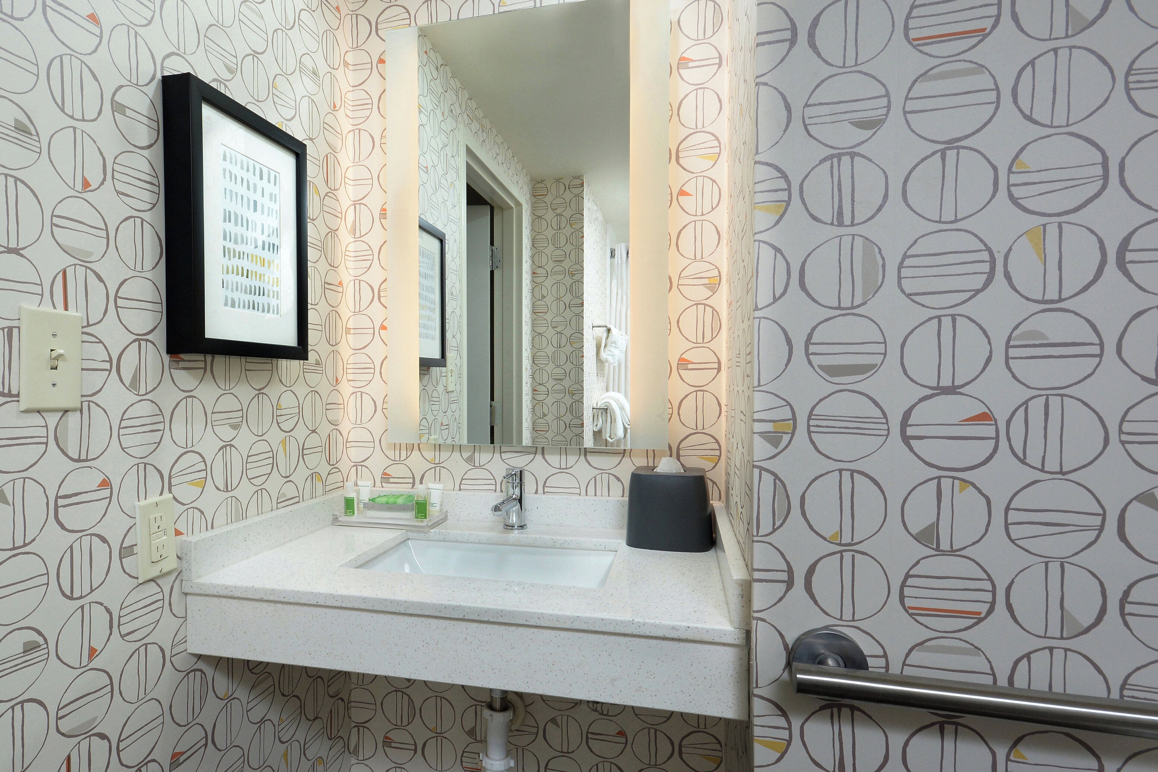A bathroom with a sink, mirror, towel dispenser, and patterned wallpaper.