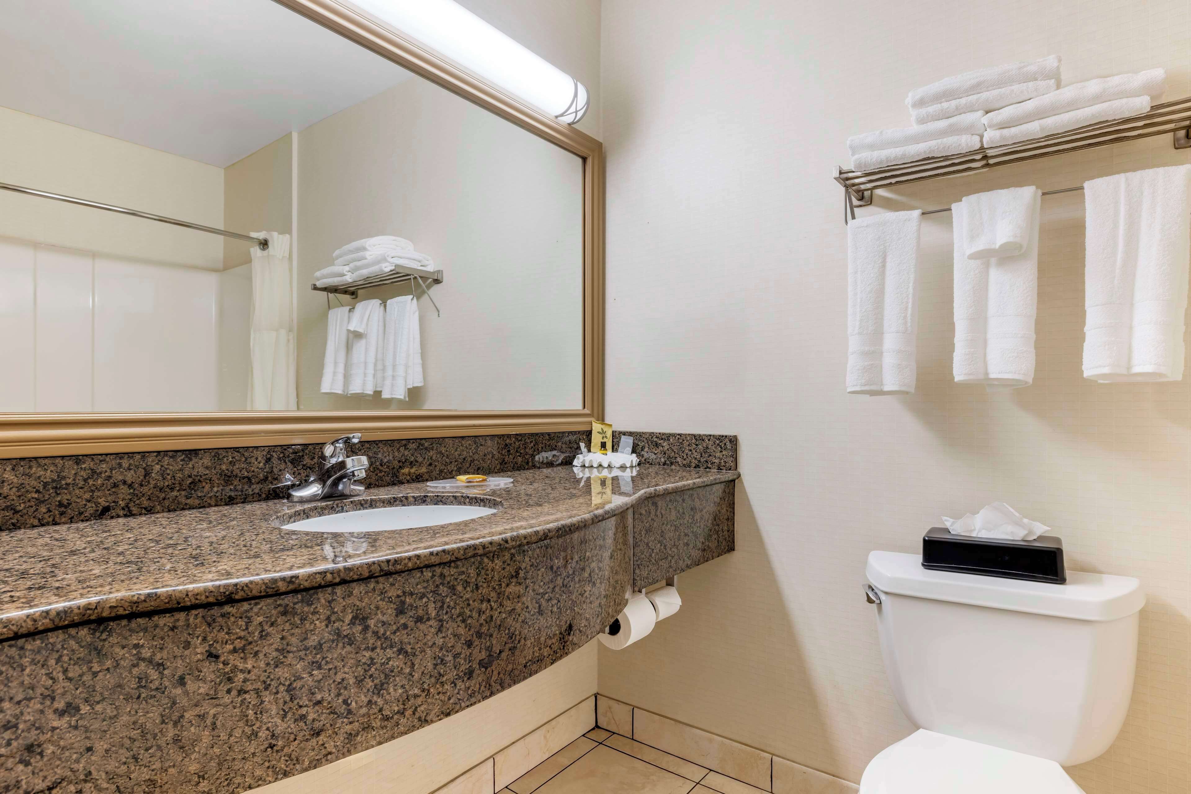 A bathroom with a granite countertop sink, a mirror, a towel rack with folded towels, a toilet, and a paper towel dispenser.