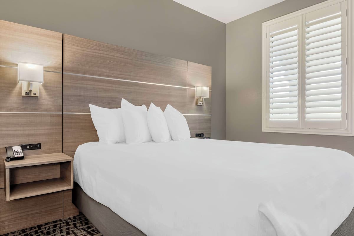 A modern hotel room with a large bed, bedside lamps, a phone on a nightstand, and a window with blinds.