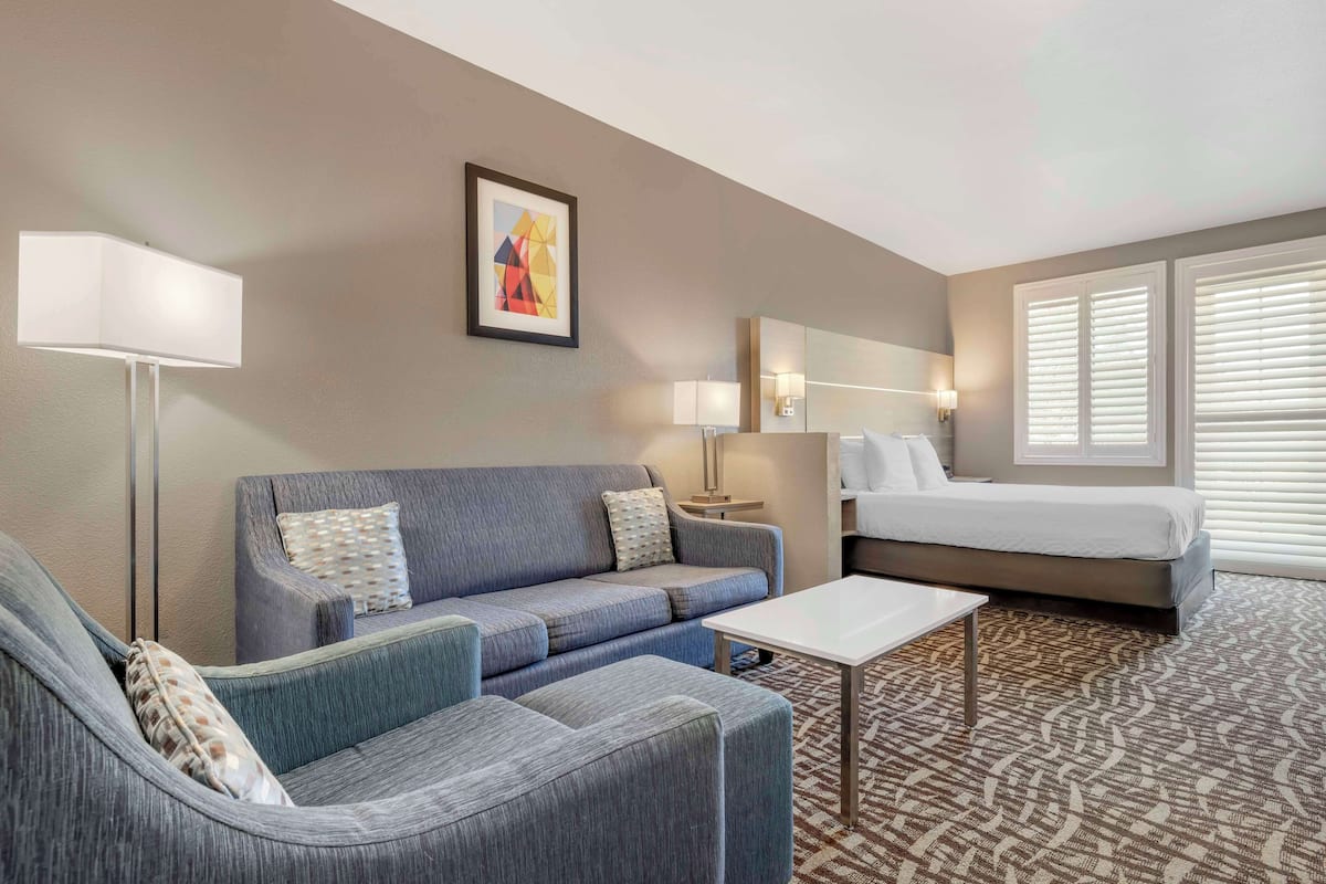 A hotel room with a grey sofa, a white coffee table, a bed with white linens, a large window with blinds, and a painting on the wall.