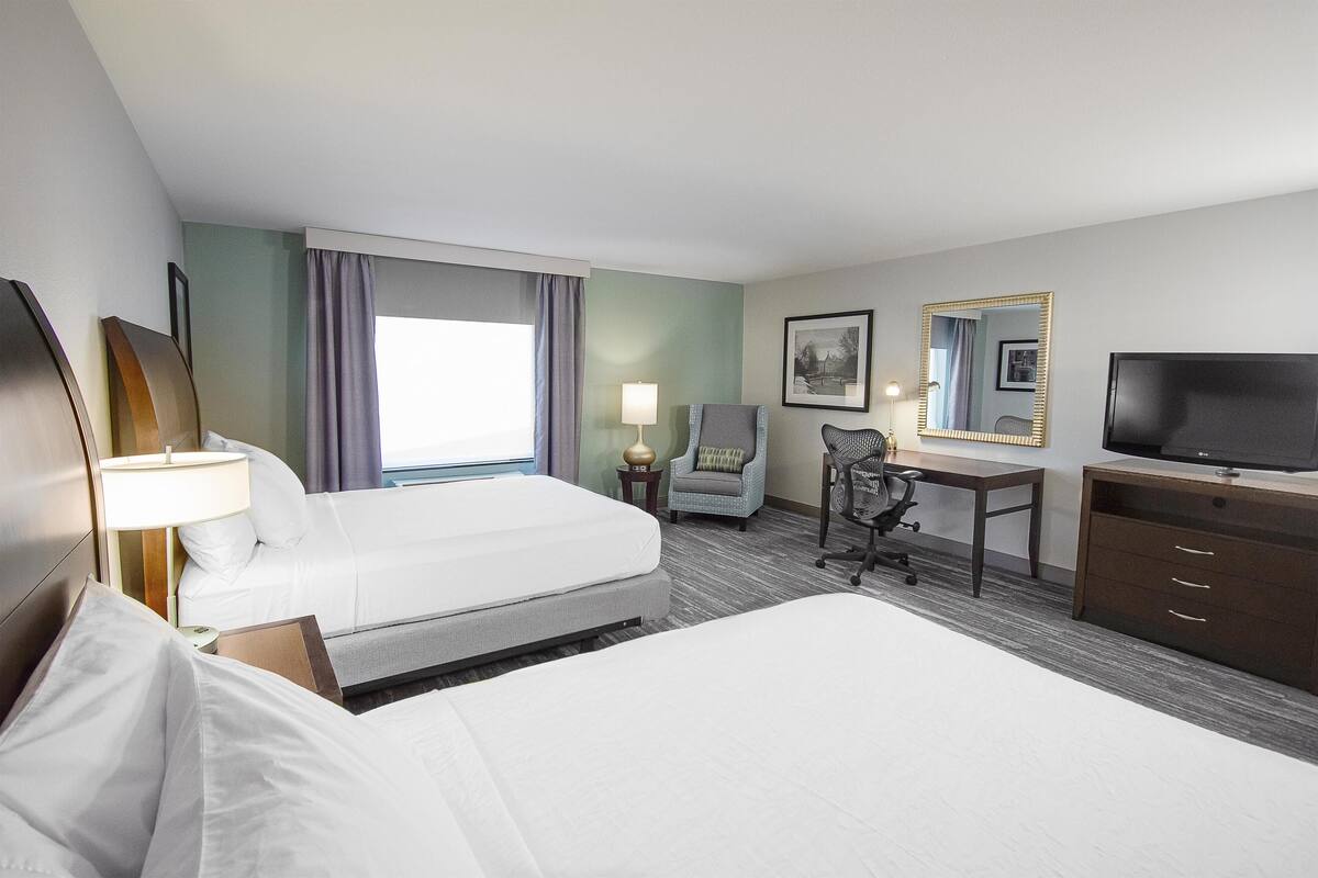 A hotel room with a large bed, a desk, a chair, a TV, and a lamp.