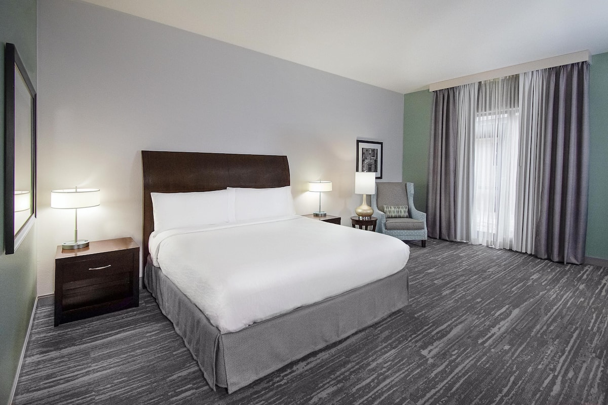 A hotel room with a large bed, two bedside tables with lamps, a chair, a framed picture on the wall, and a window with curtains.