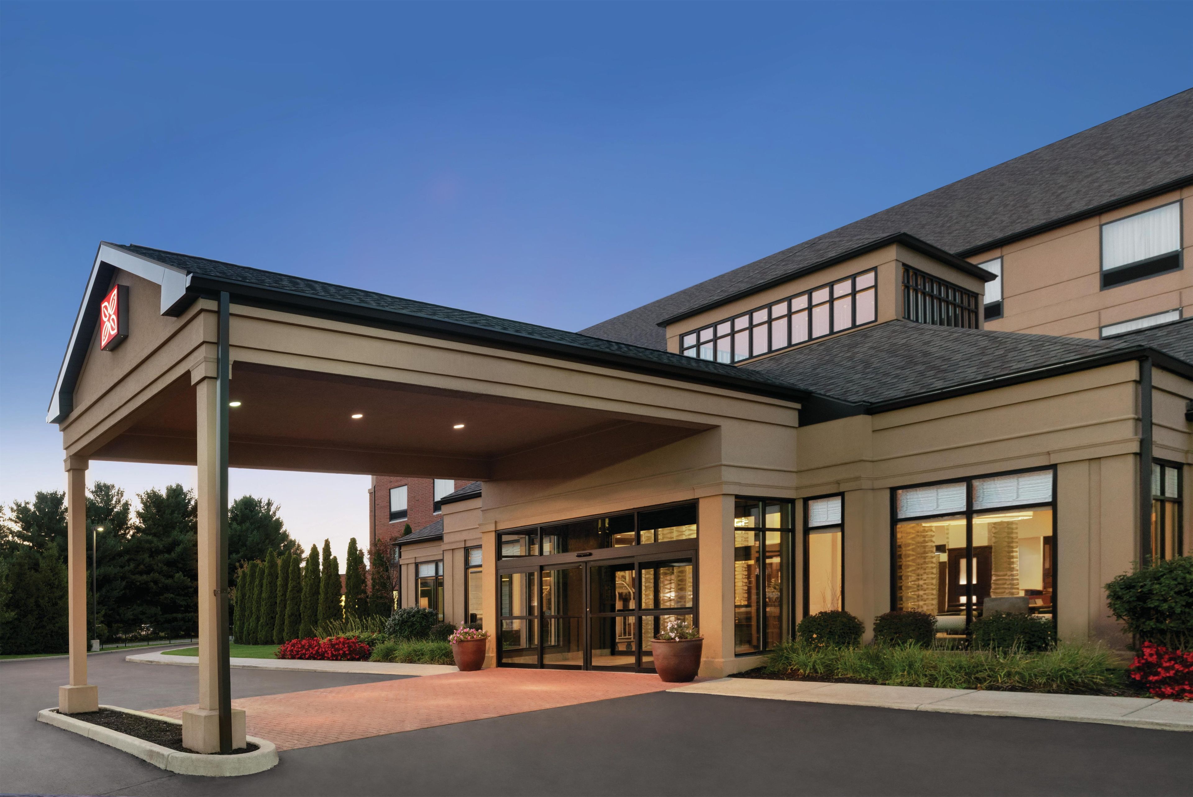 A hotel entrance with a covered walkway, large windows, and a landscaped area with potted plants.