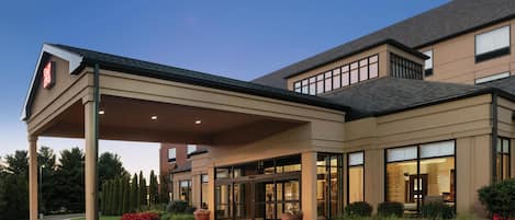 A hotel entrance with a covered walkway, large windows, and a landscaped area with potted plants.