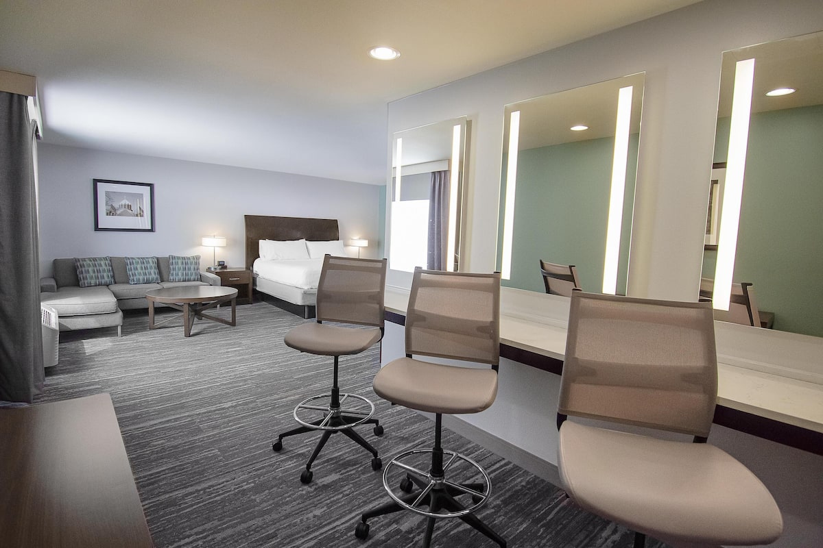 A hotel room with a bed, a sofa, two office chairs, a desk, a mirror, and a framed picture on the wall.