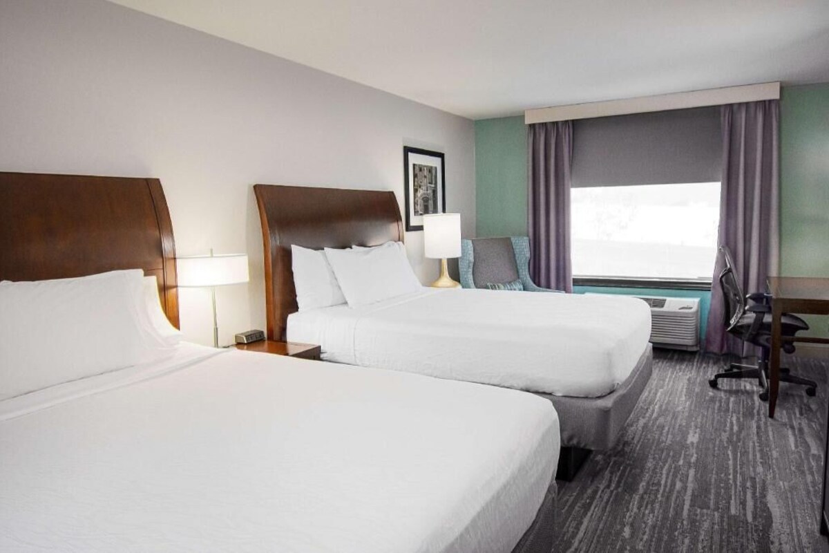 A hotel room with two beds, a desk, a chair, an air conditioning unit, and a window with curtains.