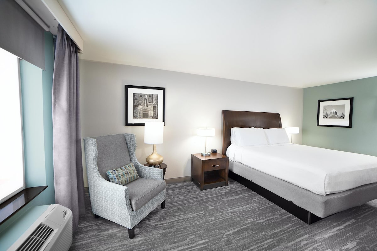 A hotel room with a bed, a chair, a nightstand with a lamp, and a window with curtains.