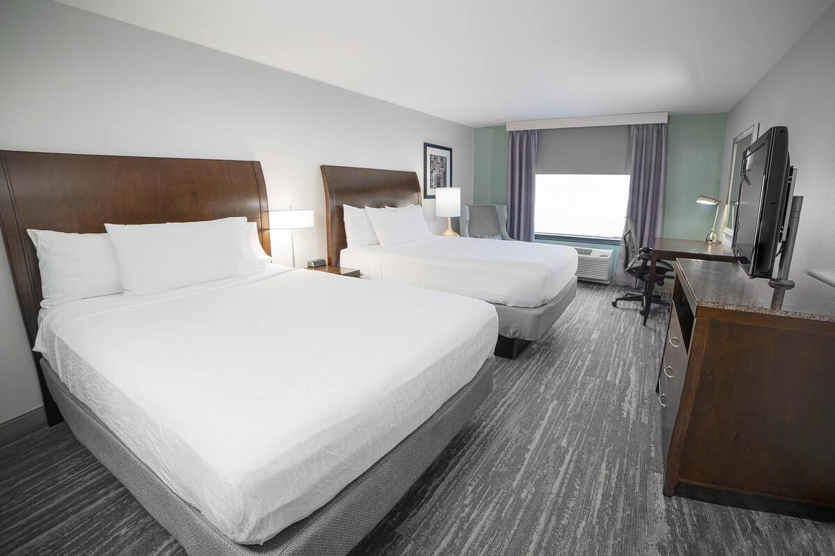 A hotel room with two beds, a desk, a chair, a TV, and a window with curtains.