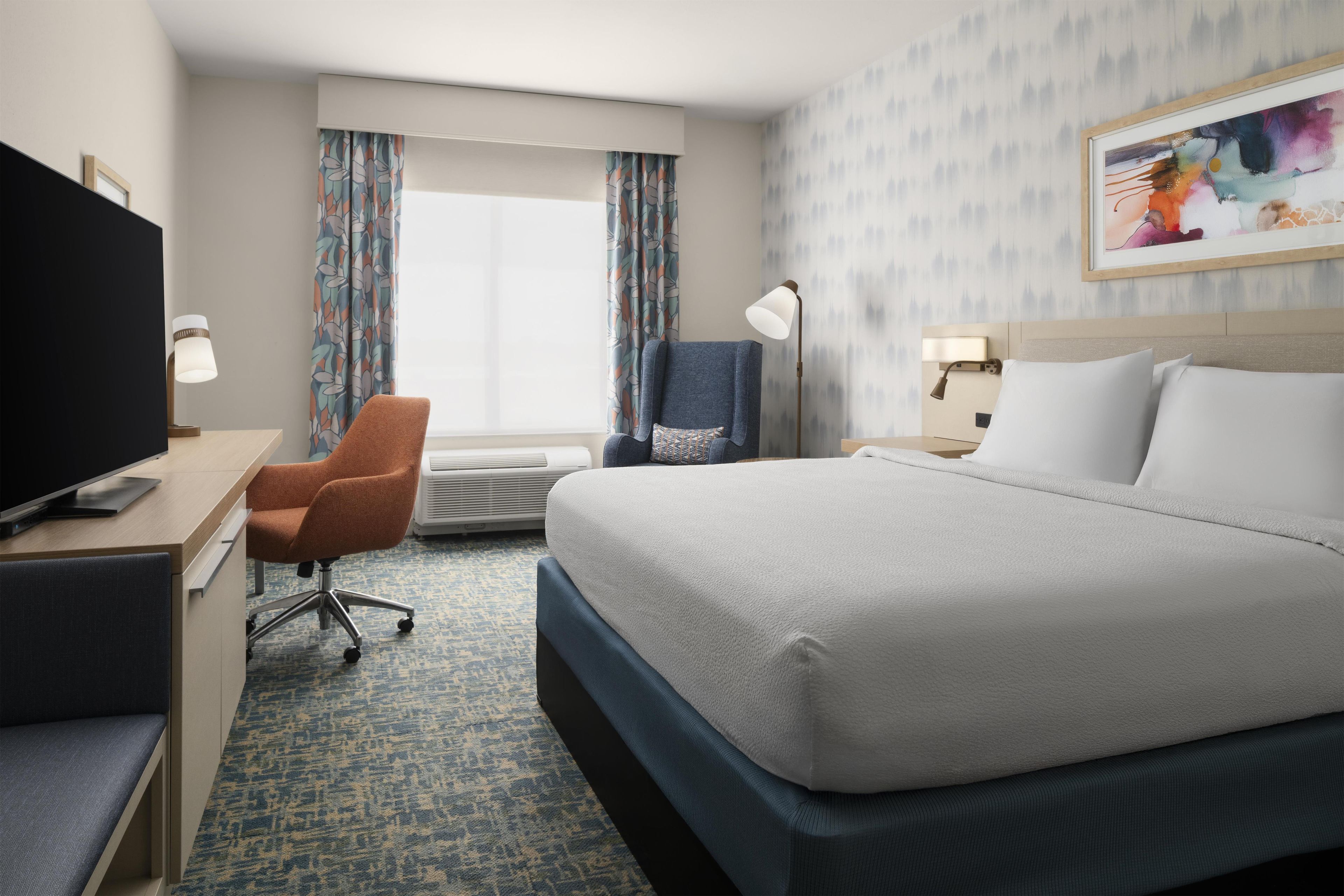 A hotel room with a bed, a desk with a television, a chair, a desk lamp, and a window with curtains.