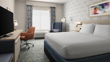 A hotel room with a bed, a desk with a television, a chair, a desk lamp, and a window with curtains.