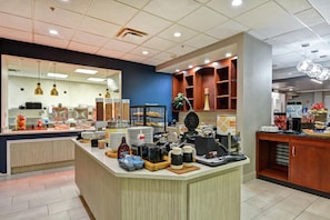 A modern cafe interior with a central counter, various food and drink dispensers, and a seating area with a wooden cabinet.