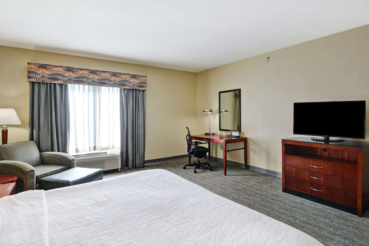 Ein Hotelzimmer mit einem großen Bett, einem Schreibtisch mit Stuhl, einem Fernseher und einem Fenster mit Vorhängen.
