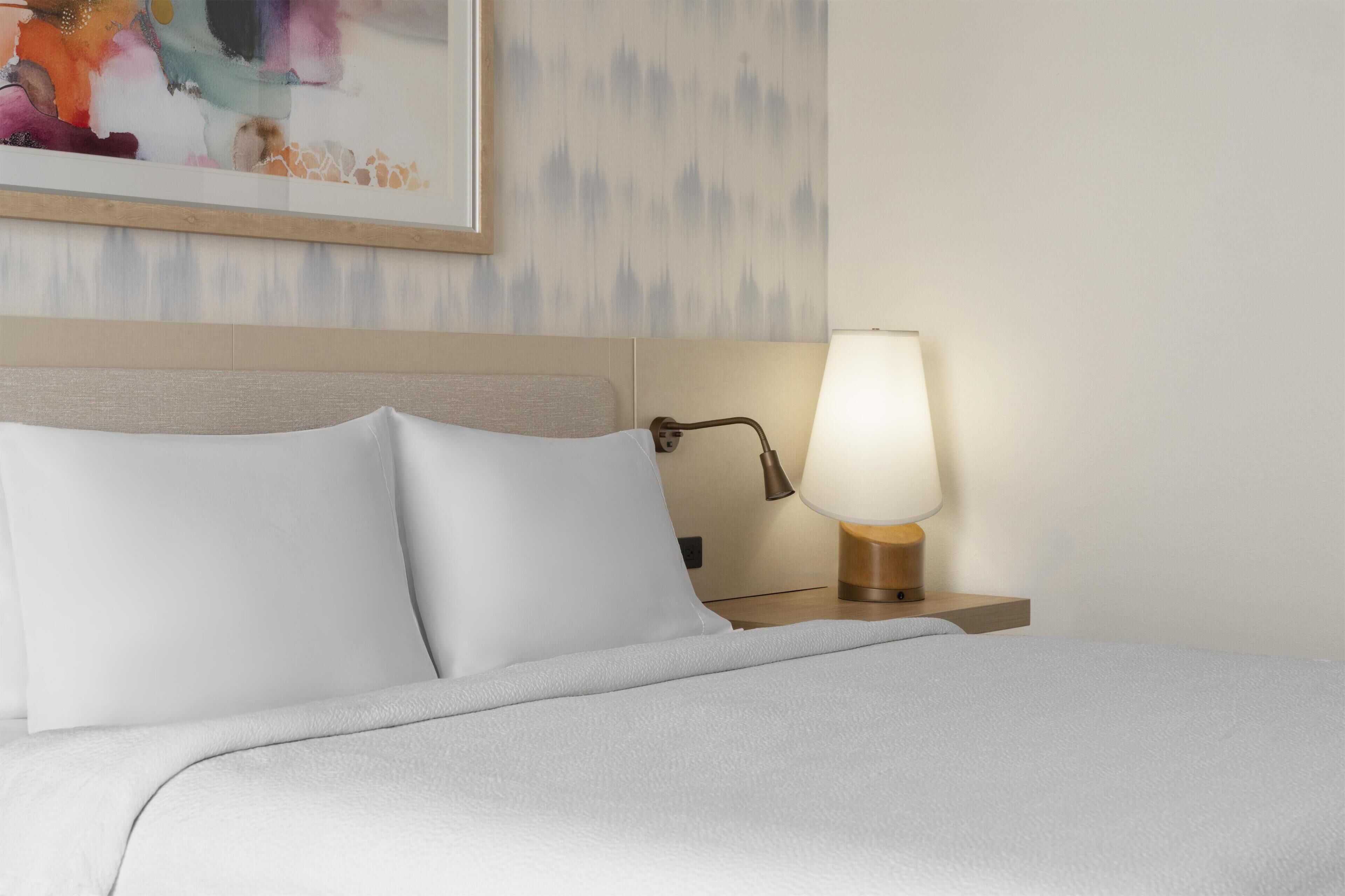 A neatly made bed with white linens and pillows, a bedside lamp, and a wall-mounted artwork.