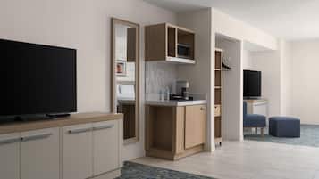 A modern hotel room with a flat-screen TV, a kitchenette, a seating area, and a large mirror.