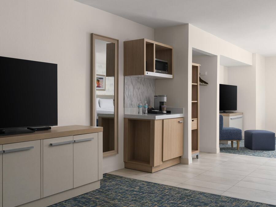 A modern hotel room with a flat-screen TV, a kitchenette, a seating area, and a large mirror.