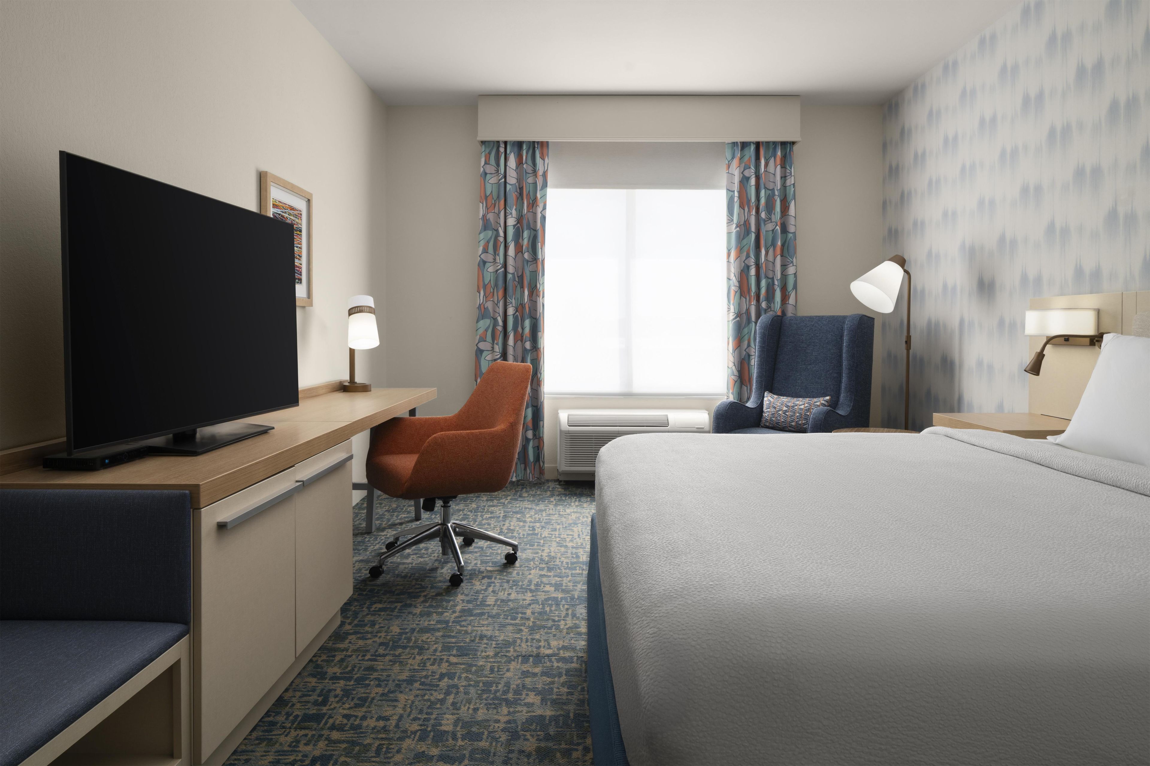 A hotel room with a bed, a desk with a flat-screen TV, a chair, a desk lamp, and a window with curtains.