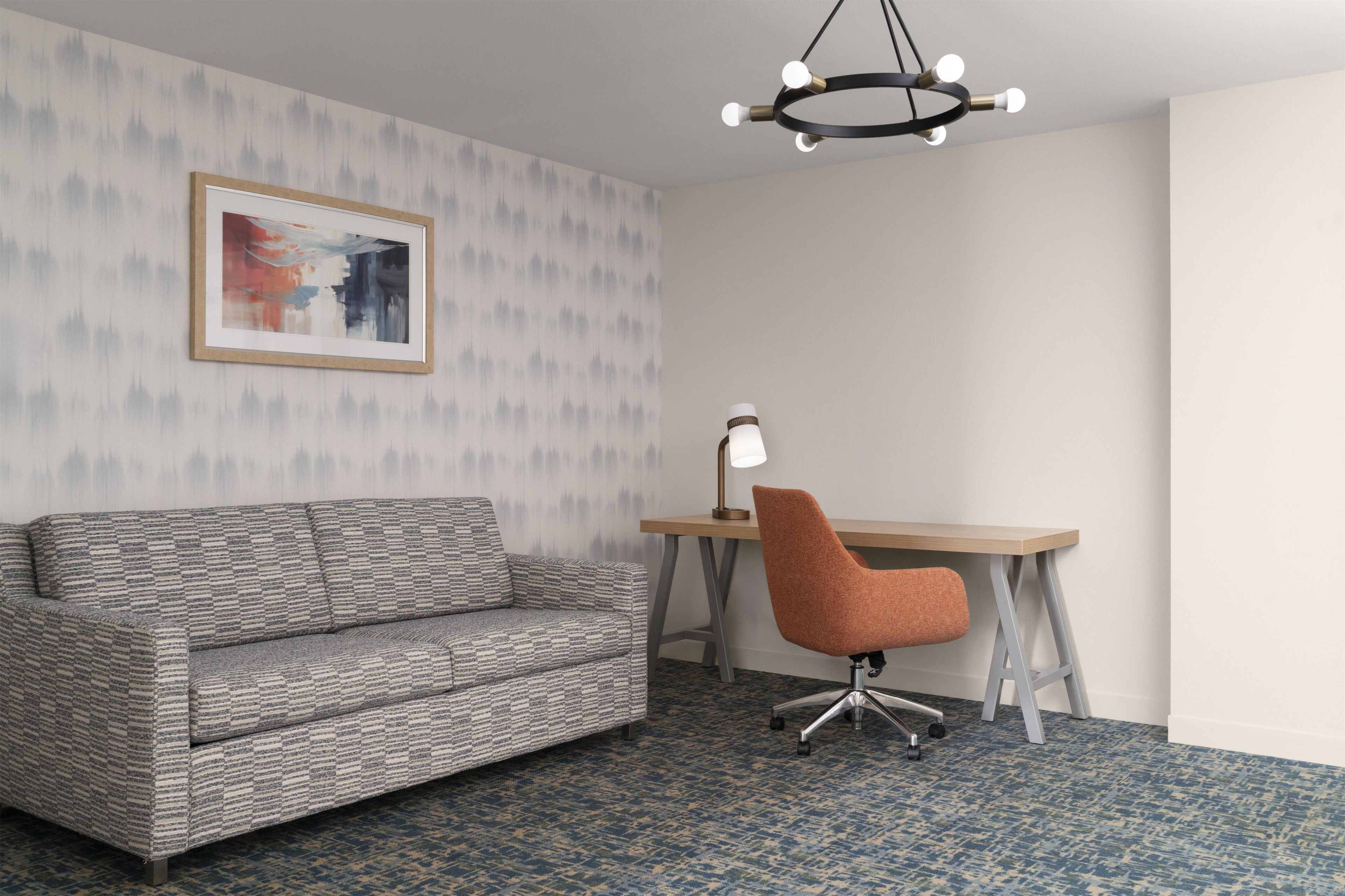 Une pièce avec un canapé à motifs, un bureau avec une chaise, une lampe de bureau et un tableau encadré au mur.