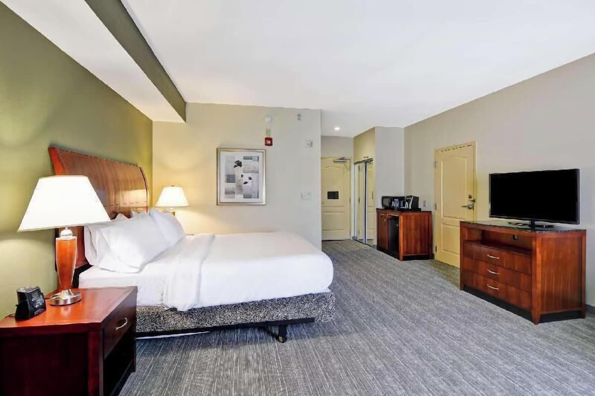 Ein Hotelzimmer mit einem Bett, zwei Nachttischlampen, einem Fernseher, einer Holzkommode und einem gerahmten Bild an der Wand.