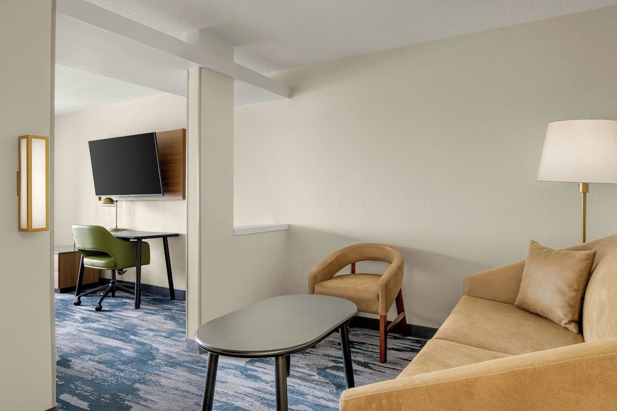 A hotel room with a sofa, a small table, a green chair, a TV mounted on the wall, and a lamp.