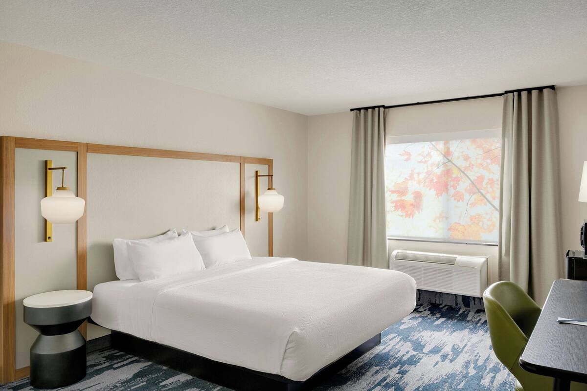 A hotel room with a large bed, two bedside lamps, a window with curtains, a desk with a chair, and a wall-mounted air conditioner.