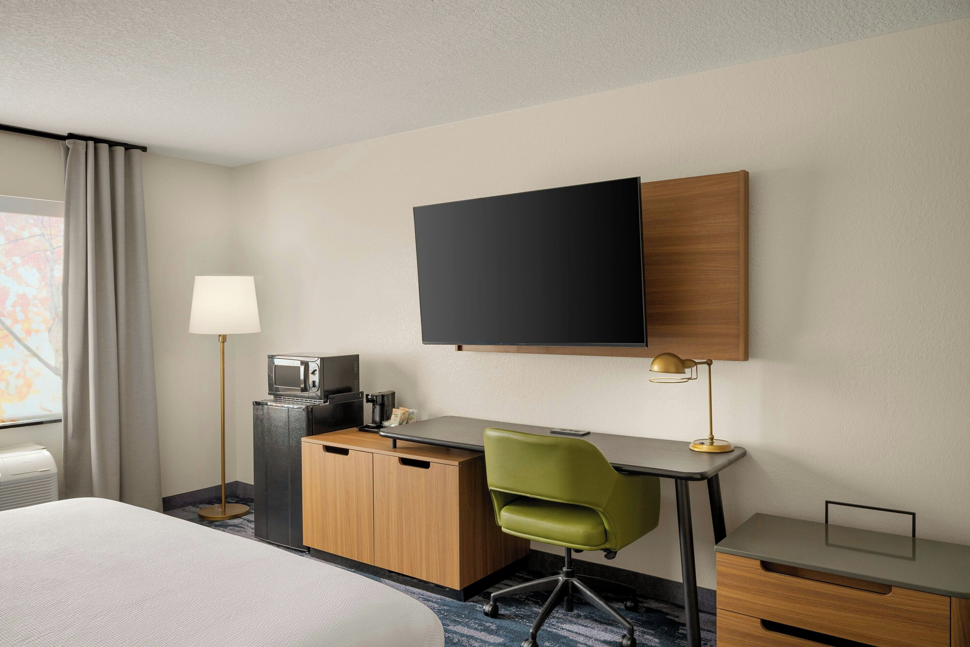 A hotel room with a bed, a desk with a chair, a television, a desk lamp, a microwave, and a minibar.