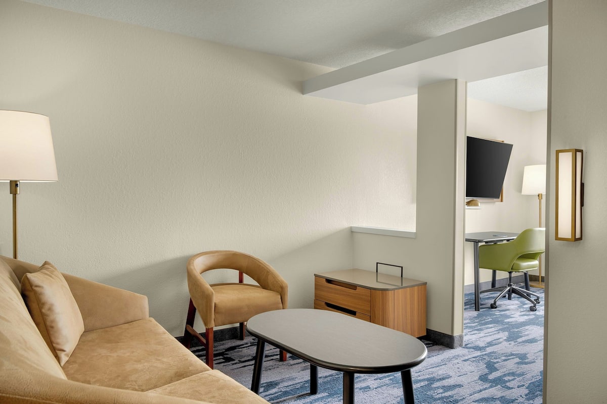 A hotel room with a beige sofa, a small table, a green chair, a wooden desk, and a TV on the wall.