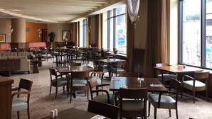 A spacious hotel dining area with multiple tables and chairs, large windows offering views of the street, and hanging pendant lights.