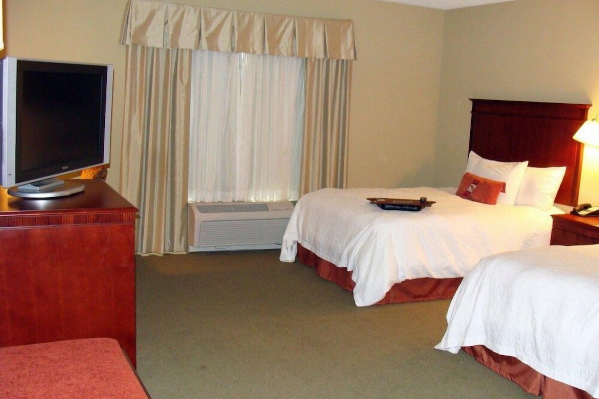 Quarto de hotel com televisão sobre uma cômoda de madeira, cama com roupa de cama branca, janela com cortinas e mesa de cabeceira com abajur.