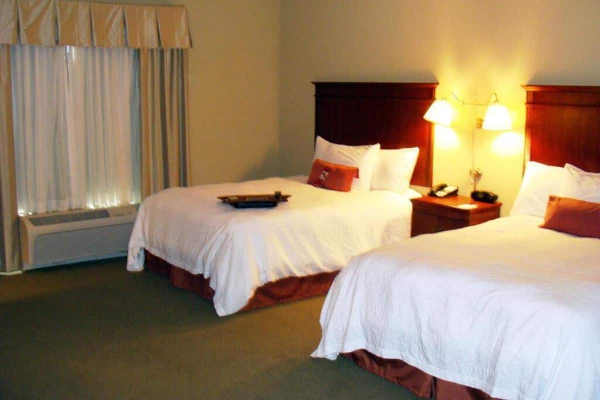 A hotel room with two beds, a window with curtains, a bedside table with a lamp, and an air conditioning unit.
