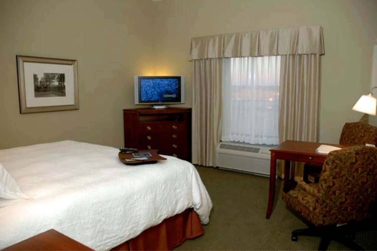 Une chambre d’hôtel avec un lit, une télévision, un bureau, une chaise, un tableau encadré au mur et une fenêtre avec des rideaux.