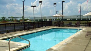 A rectangular swimming pool with a metal safety railing, surrounded by a concrete deck. There are outdoor chairs and umbrellas, and a fence enclosing the area.