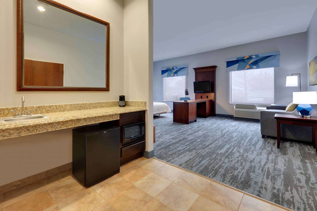 A hotel room with a kitchen area, a bed, a TV, and a large window.
