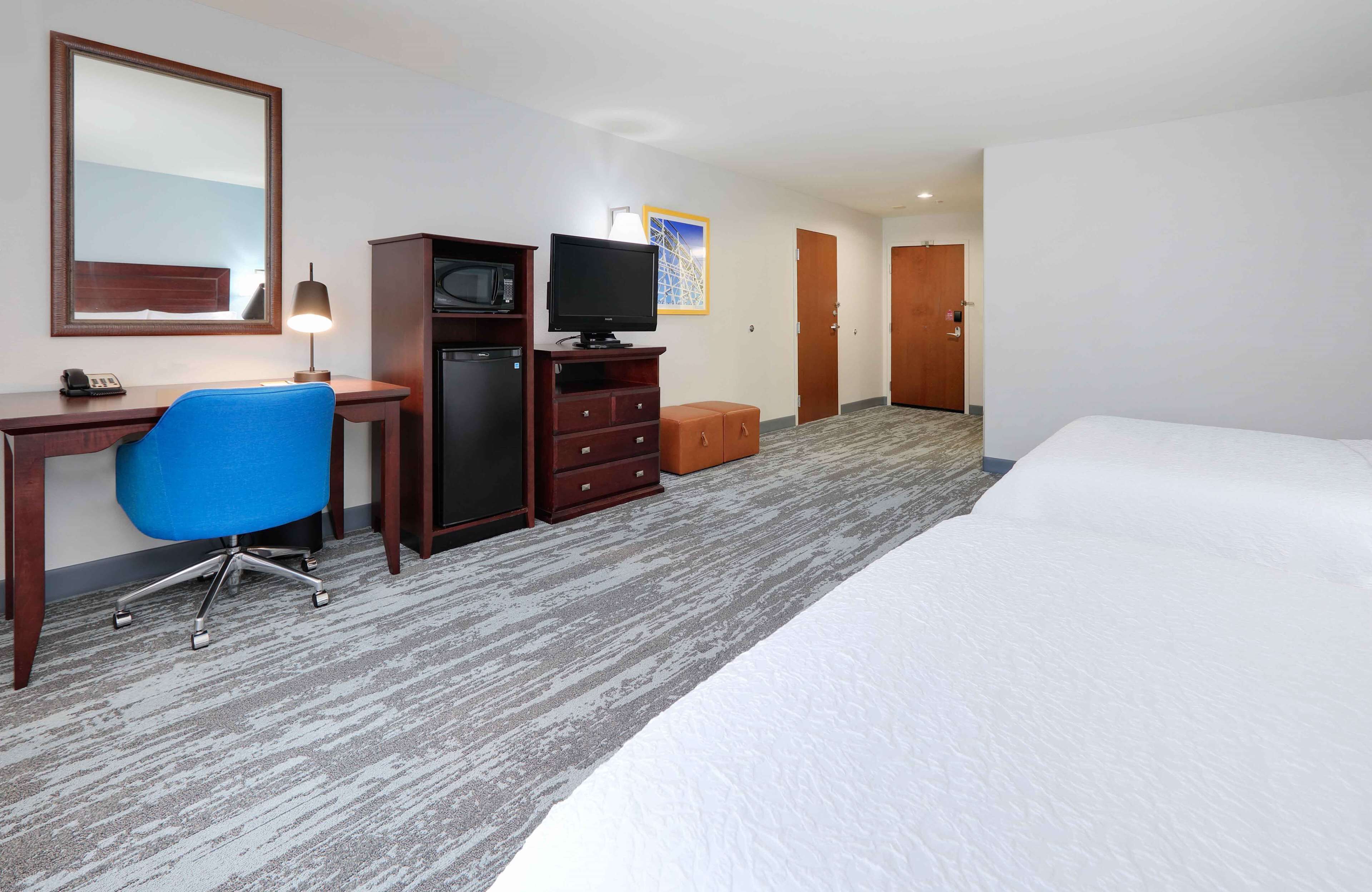 A hotel room with a bed, a desk with a computer, a chair, a TV, a microwave, and a small cabinet.