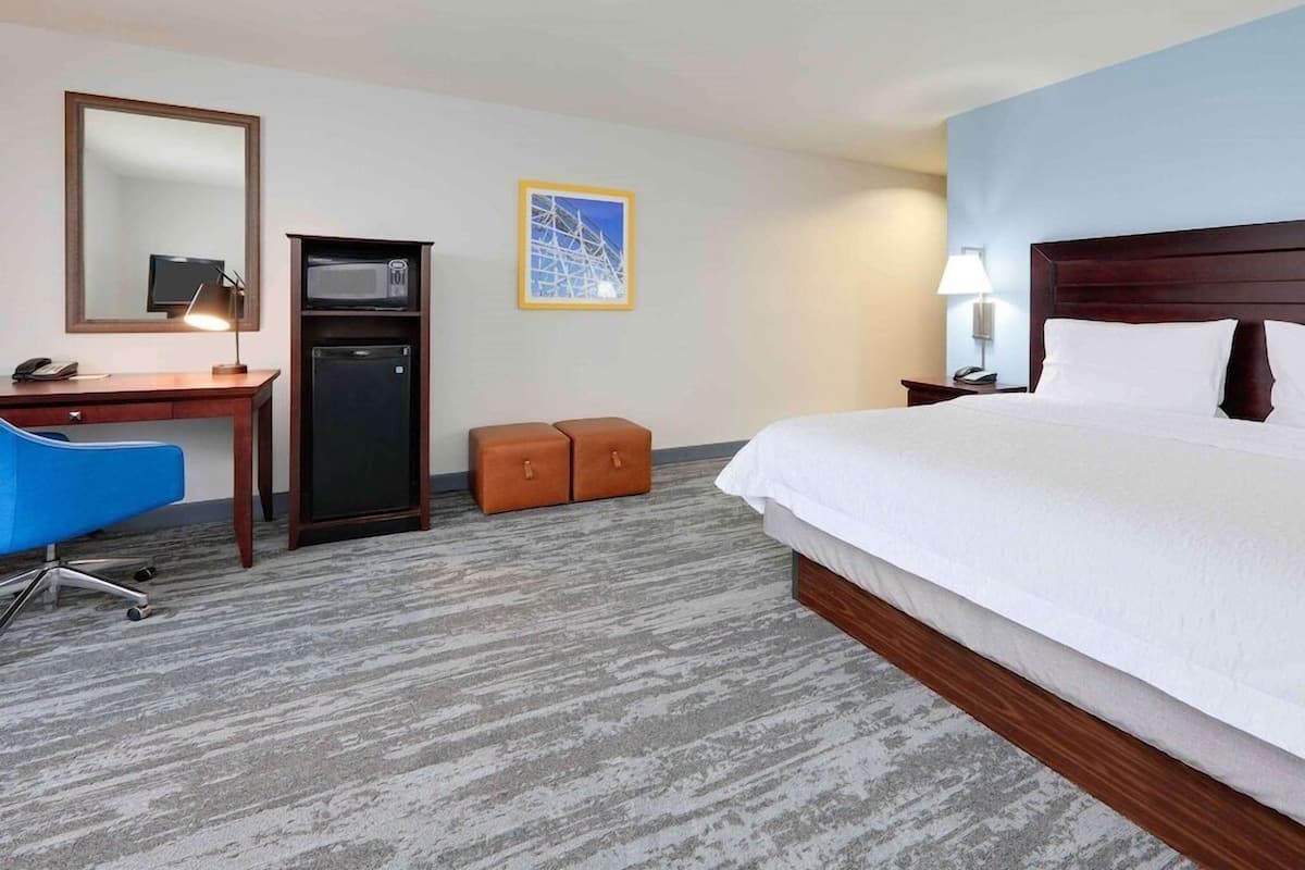 A hotel room with a bed, desk, chair, microwave, refrigerator, and a framed picture on the wall.