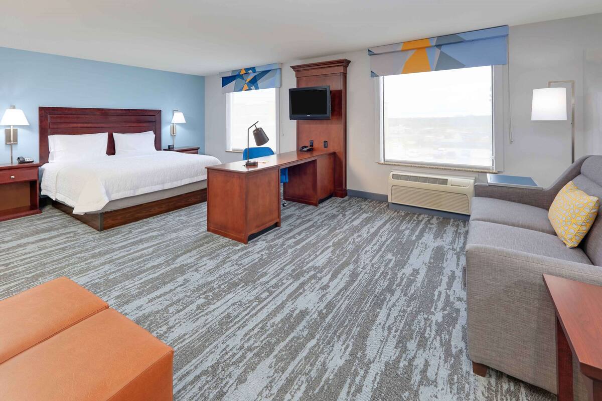 A hotel room with a bed, desk, TV, sofa, and large windows.
