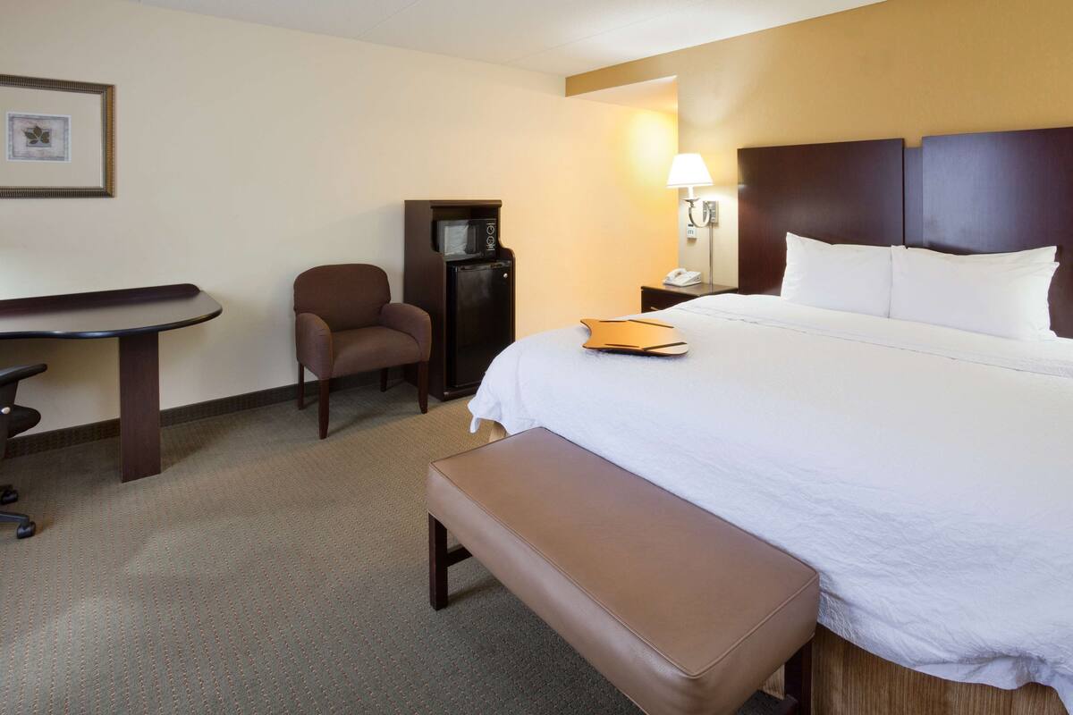 A hotel room with a bed, a desk, a chair, a small table, and a bench.