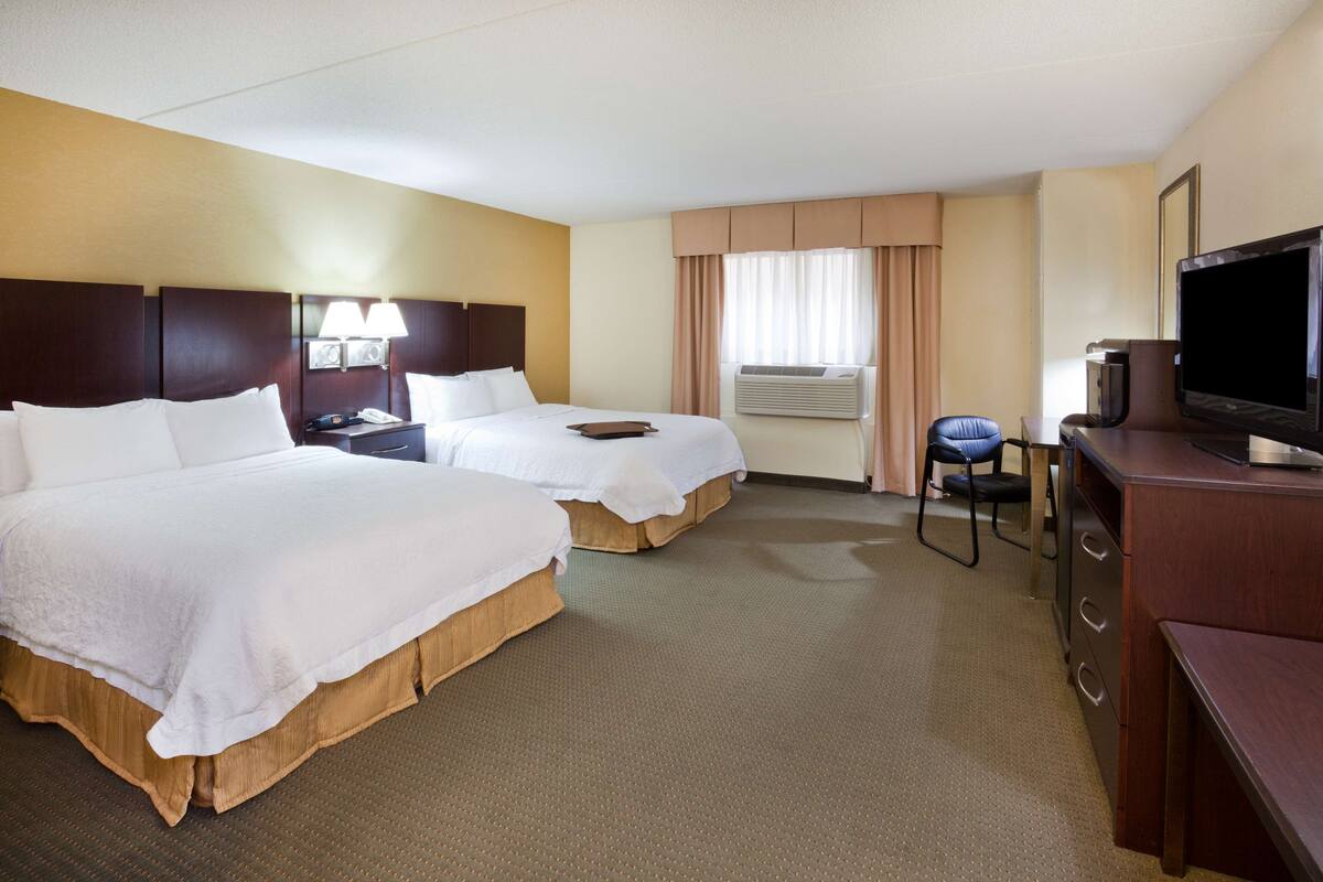 A hotel room with two beds, a desk with a television, and an air conditioning unit.