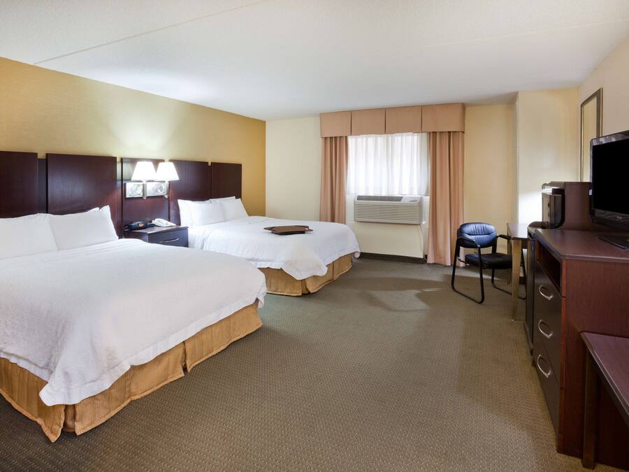 A hotel room with two beds, a desk with a television, and an air conditioning unit.