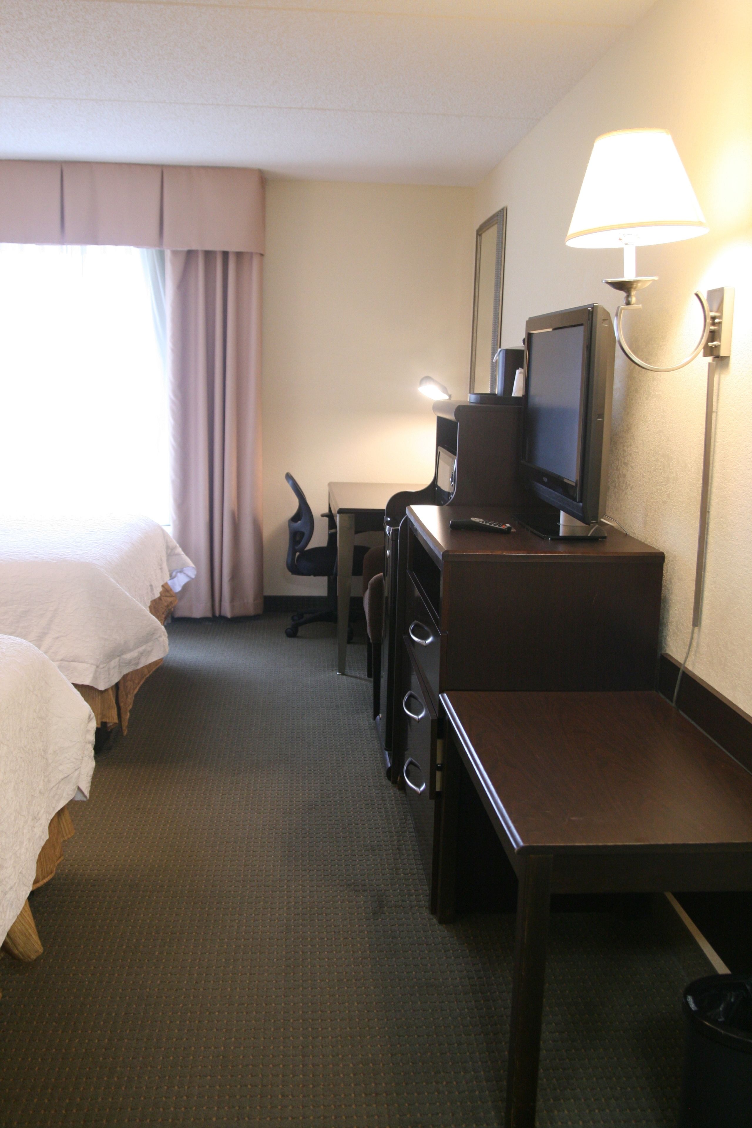 A hotel room with a bed, a desk with a television, a chair, and a lamp.